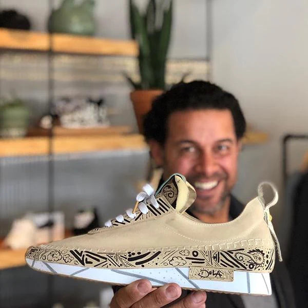 A person with a large smile is holding up a single, custom-designed ComunityMade sneaker. The shoe is a light tan leather with intricate black line drawings covering the side and a small section on the tongue. The drawings feature various abstract shapes, patterns, and a small fish icon. The shoe has white laces and a thick white sole with a subtle geometric pattern and a light gray stripe. The heel has a small white loop.
