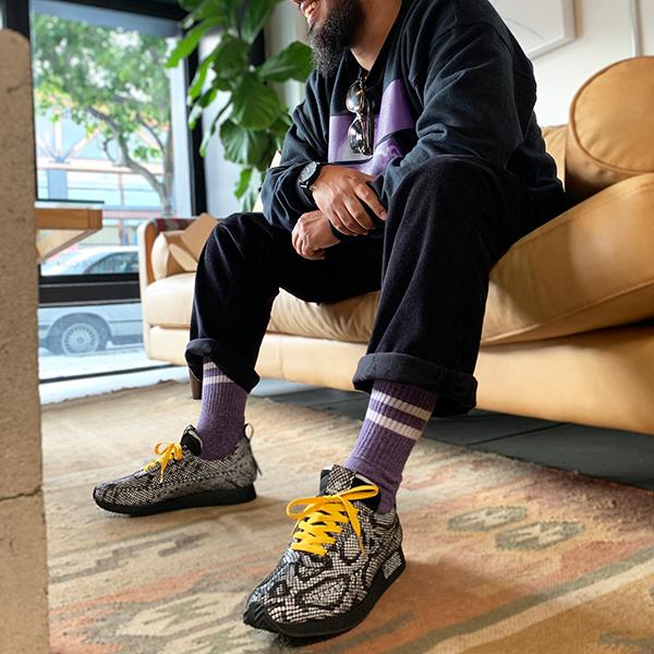 A low-angle shot of a man's lower body, sitting on a light brown leather couch. He is wearing dark pants with cuffed hems, showing off purple and white striped socks and a pair of distinctive sneakers. The sneakers have a black and white snakeskin-like pattern, with thick black soles and bright yellow laces. He is sitting in a casual, relaxed pose on an ornate patterned rug.