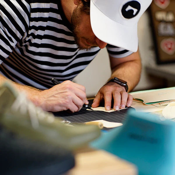 A man wearing a striped shirt and a white baseball cap with a ComunityMade logo is focused on a task at a workbench. He is using a small tool, possibly a pen or an awl, to work on a piece of light-colored material placed on a black cutting mat. Part of a shoe is visible in the blurry foreground.