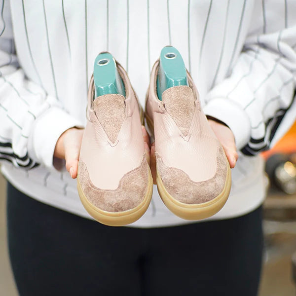 A person is holding up a pair of finished, light-colored shoes, likely handcrafted. The shoes are a soft pinkish-tan leather with a darker, textured suede material on the toes. They have tan rubber soles and a low-cut, slip-on style. The person is wearing a white and black striped shirt and the shoes contain light blue shoe trees to help them keep their shape.