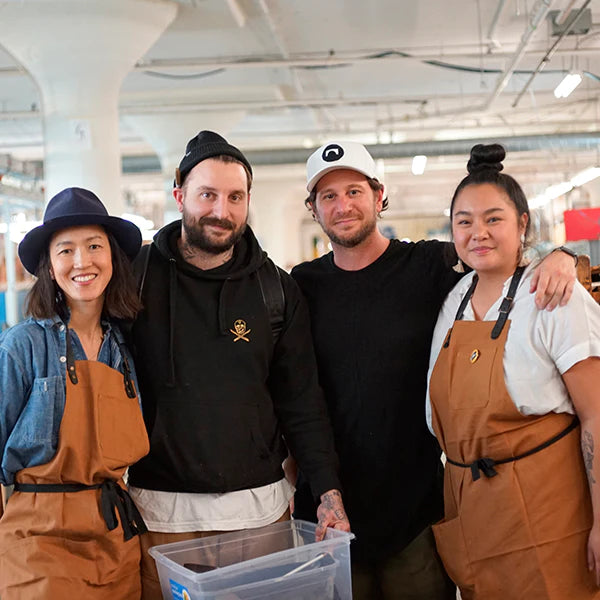 A group portrait of four people standing together in a workshop or studio setting. From left to right, a woman in a navy hat and brown apron smiles at the camera. Next to her, a man with a beard and tattoos, wearing a black beanie and a black hoodie, has his arm around the shoulder of another man. The second man is wearing a white baseball cap and a black t-shirt and has a tattoo on his left forearm. On the far right, a woman with her hair in a bun and wearing a brown apron smiles..