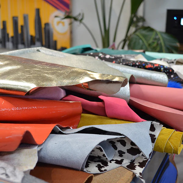 A close-up image of a pile of various leather and fabric swatches, for shoe-making The materials are stacked on top of each other, displaying a wide range of colors and textures. Visible materials include a shiny gold piece, a bright orange piece, a pink one, and a piece with a black-and-white cow print. In the blurry background, a graphic of a city skyline is visible.