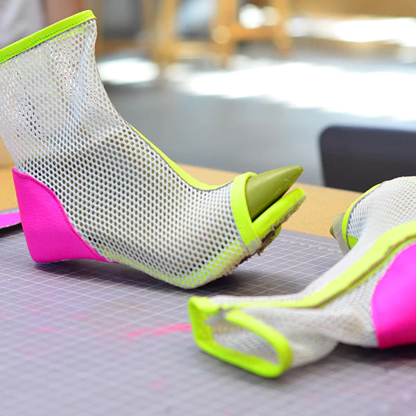 A close-up of a pair of unique, unfinished shoes with a vibrant, modern design. The shoes are made of a white mesh material with neon lime green trim and a hot pink accent on the heel. One shoe is standing upright, showing its pointed green toe and a wedge heel. The other shoe is lying on its side. Both are placed on a gray cutting mat on a wooden table.