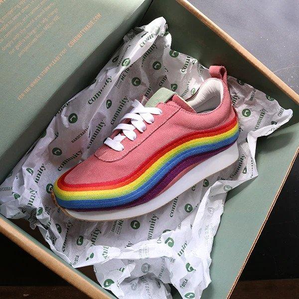 
A single pink sneaker with white laces is nestled in a green shoe box, resting on crinkled white tissue paper printed with a repeating "community" logo. The sneaker features a thick platform sole with a rainbow stripe design running along the side, from top to bottom. The tongue of the shoe is a light green color, and there is a red loop on the back of the heel. The box lid is visible and has a "comunitymade" logo on the inside.
