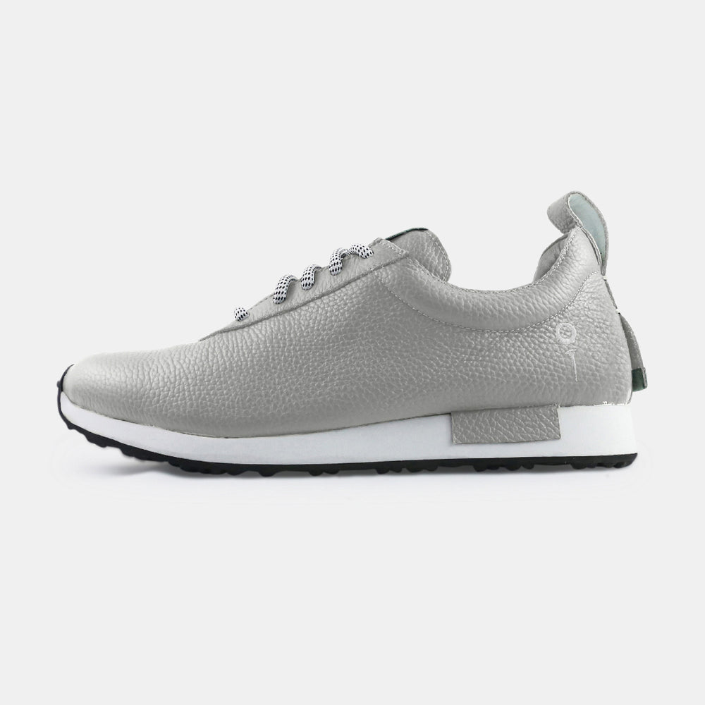 
                      
                        A gray, near silver pebbled leather ComunityMade sneaker with a thick white sole. The shoe has black laces with white flecks, a small gray leather logo tag on the side of the heel, and a mint green pull tab at the back.
                      
                    