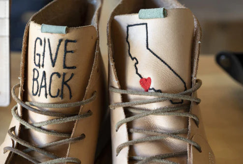A close-up shot of a pair of light tan leather shoes with laces. The tongue of the left shoe has "GIVE BACK" written on it in black marker. The tongue of the right shoe has an outline of the state of California with a small red heart stitched inside. Both shoes have a light teal-colored loop at the top of the tongue.