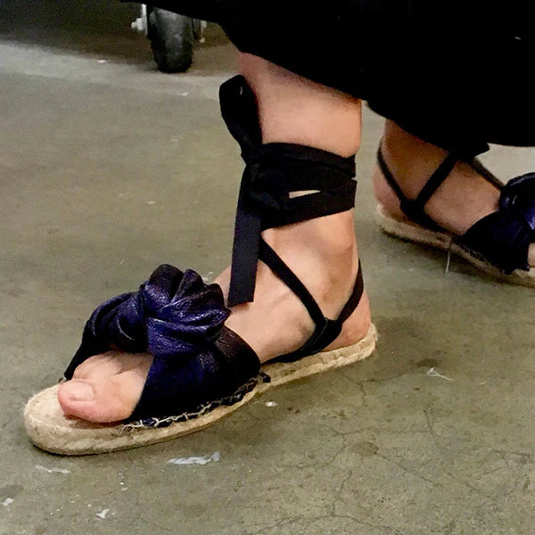 A low-angle, close-up shot of a person's feet wearing a pair of stylish espadrilles. The shoes have a flat, woven jute sole and black fabric straps. The straps wrap around the ankle and are tied in a bow, and another large bow-like detail is on the top of the foot. The person is standing on a gray, concrete floor.