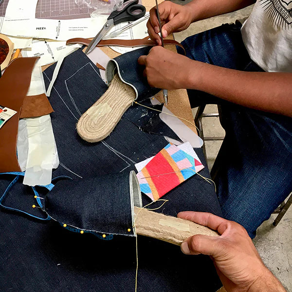 A close-up view of two people's hands working on a handcrafted shoe. One person is using a needle and thread to stitch a dark blue denim fabric to a shoe sole made of rope or jute. The other person's hands are holding a similar sole and a piece of brown leather. Various shoe-making tools and materials, including scissors, a heel pattern, and other pieces of fabric and paper, are scattered across the workbench.