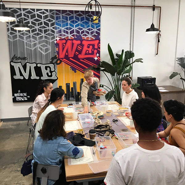 A group of seven people, primarily women, is gathered around a large table in what appears to be a workshop or a classroom. A young person with blonde hair, wearing a striped apron, stands at the head of the table and is holding up a shoe sole, seemingly instructing the others. The attendees are seated and focused on the person speaking. The room has a modern, creative aesthetic, with a large, colorful mural on the wall that features the words "from ME to WE" and a skyline graphic.