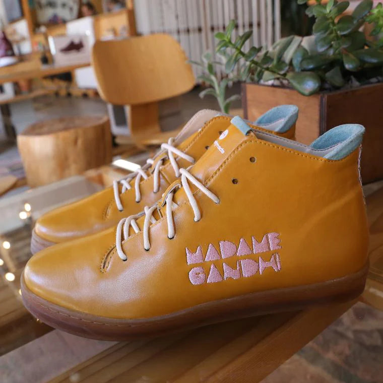 A pair of high-top shoes is sitting on a wooden surface, likely a table. The shoes are a vibrant mustard yellow or tan leather, with white laces and a light brown sole. The inside lining is a light teal color. The side of the right shoe is embroidered with the name "MADAME GANDHI" in a light pink thread. In the background, there is a blurry view of a room with furniture and plants.