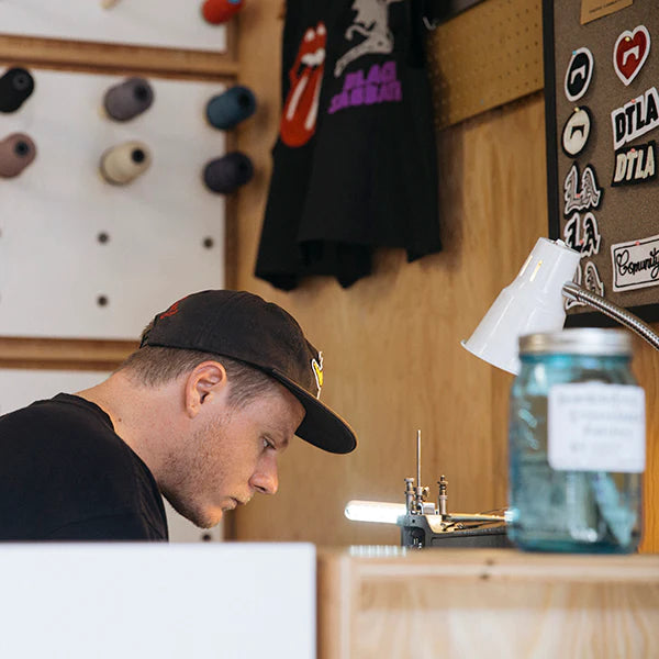 A close-up side profile of a man wearing a black baseball cap, intently focused on a task at a small machine on a workbench. A white desk lamp is illuminating the work area. In the background, there is a wooden wall with a pegboard holding spools of thread and several stickers. A black t-shirt with a "Black Sabbath" logo is hanging on the wall. A blue mason jar sits on the workbench to the right.