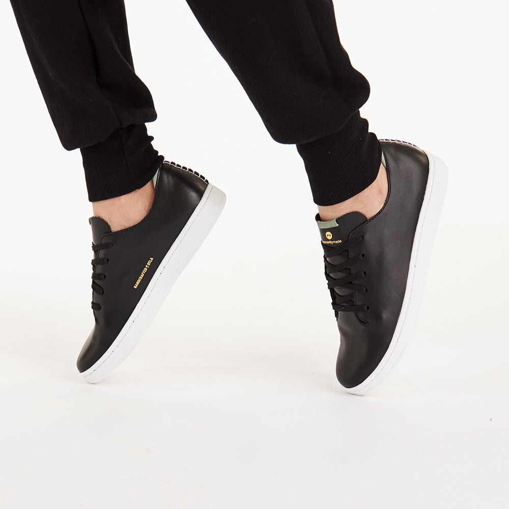 
                      
                        A picture of a person from the knee down standing on their toes while wearing a pair of black leather laceup sneakers with a white outsole and a gold print on the tongue that says COMUNITYmade with a circle logo. The person is wearing black cuffed sweatpants on a white background.
                      
                    