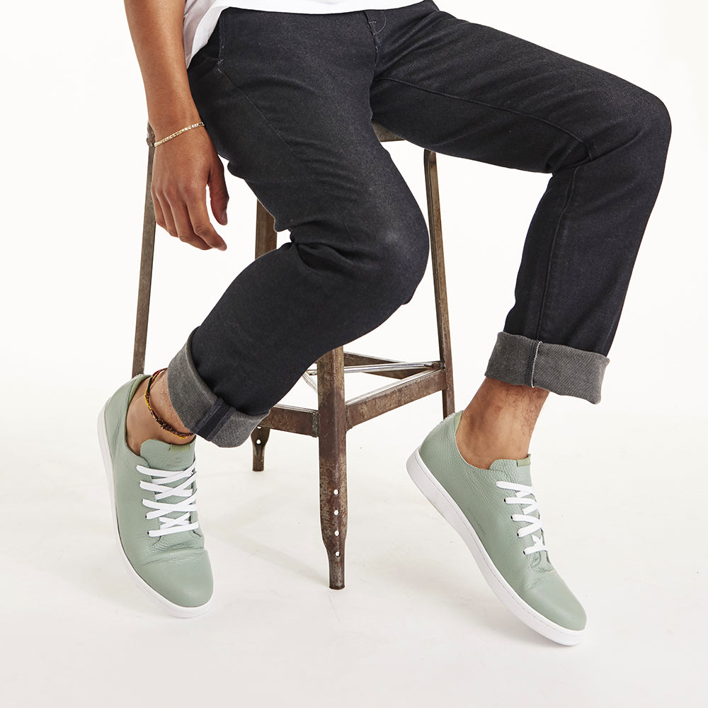 
                      
                        A picture from the waist down of a person sitting on a metal stool wearing a white t-shirt and blue jeans and a pair of light green COMUNITYmade Westsider lace-up sneakers 
                      
                    
