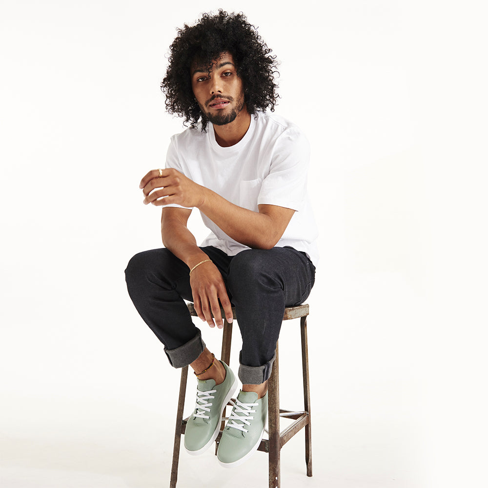 
                      
                        A picture of a male with curly black hair sitting on a metal stool with a white t-shir tand blue jeans wearing a pair of light green COMUNITYmade Westsider lace-up sneakers 
                      
                    