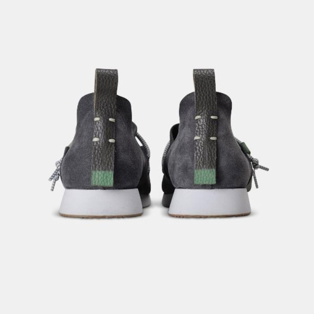 
                      
                        A back-angle, heel product shot of a pair of grey suede slip-on shoes. The shoes have thick, speckled black and white rope-like laces that are tied in a loose knot across the top of the foot. The insoles are dark gray with the word "comunity" and a circular logo printed on them in white. The shoes have a small brown leather loop on the heel. The background is a solid light gray.
                      
                    