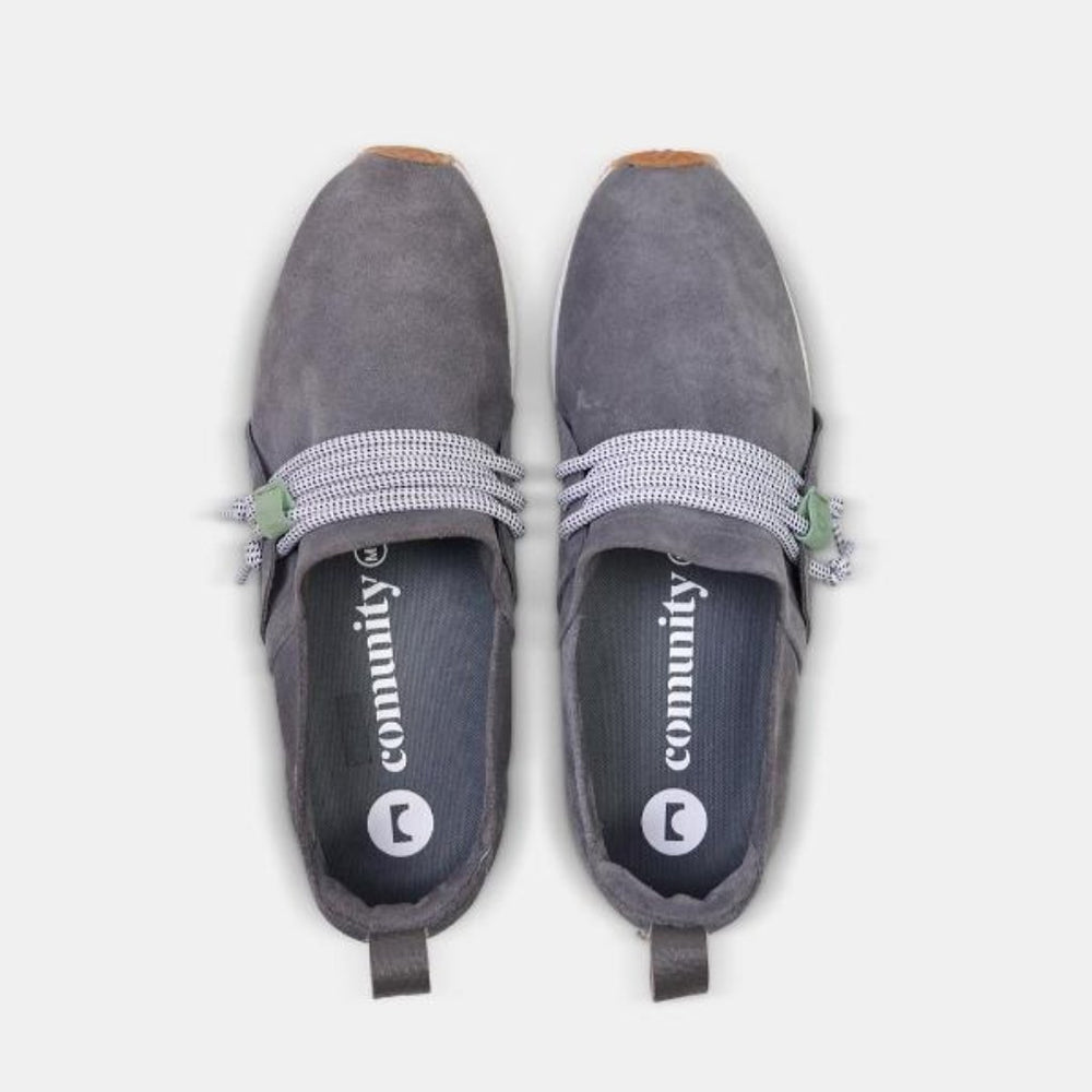 
                      
                        Mateos, A high-angle, top-down product shot of a pair of brown suede slip-on shoes. The shoes have thick, speckled black and white rope-like laces that are tied in a loose knot across the top of the foot. The insoles are dark gray with the word "comunity" and a circular logo printed on them in white. The shoes have a small brown leather loop on the heel. The background is a solid light gray.
                      
                    