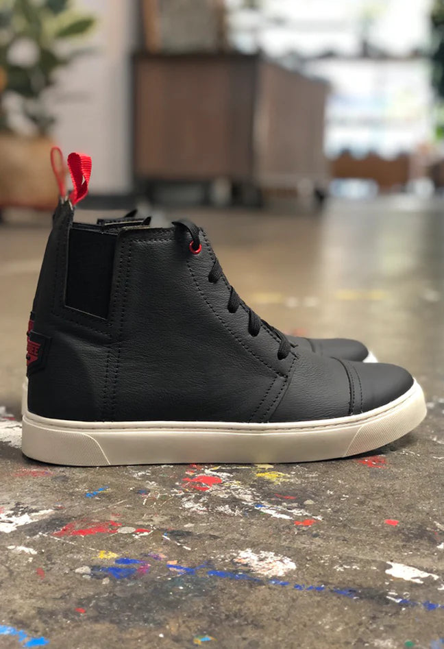 A low-angle shot of a pair of black high-top sneakers, placed on a paint-splattered concrete floor. The shoes are made of black leather with a white rubber sole. They have black laces, red stitching accents, and a red loop on the back of the heel. The shoes have a sleek, modern design, blending elements of a classic sneaker with a boot.