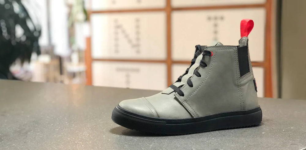 A single high-top shoe, made of smooth gray leather, is placed on a gray countertop. The shoe has black laces and a thick black sole. A red loop is attached to the back of the heel. The shoe combines the look of a sneaker and a boot, with a clean, modern design. In the blurry background, there are white boxes with black letters on them.