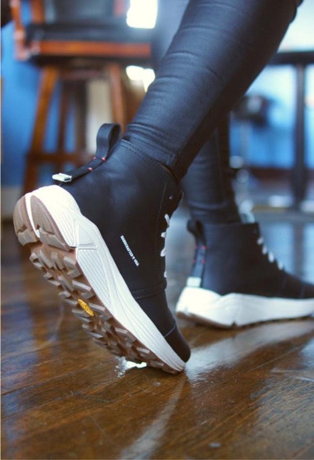 Below the knee photo of a person wearing black tights walking away wearing black mid-cut sneakers and a white midsole with a gum rubber outsole