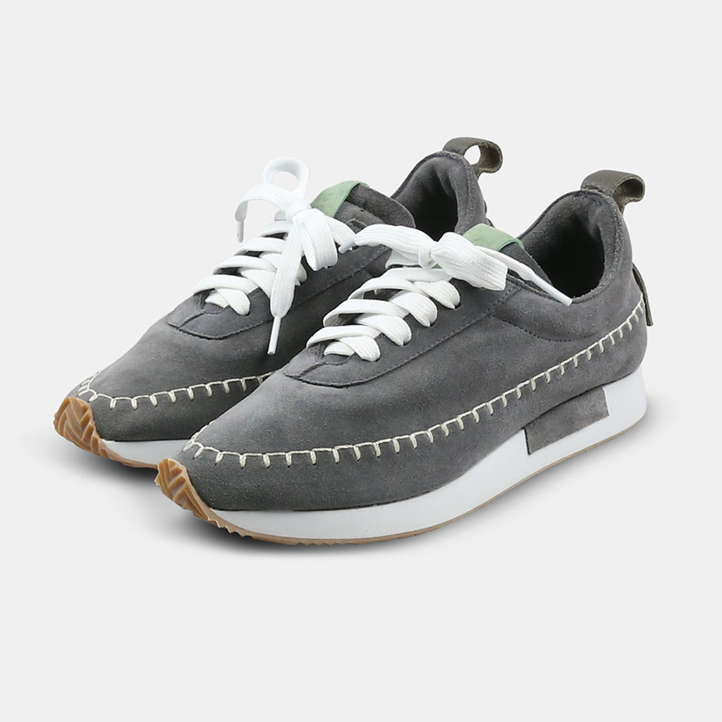 A quarter profile shot of a pair of grey suede Comunitymade lace-up shoes. The shoe is made of a grey suede with a green tongue accent.  It has flat white laces and white accent stitching around the entire shoe, plus a grey heel pull tab. It has a flat white midsole with a gum-rubber colored outsole.