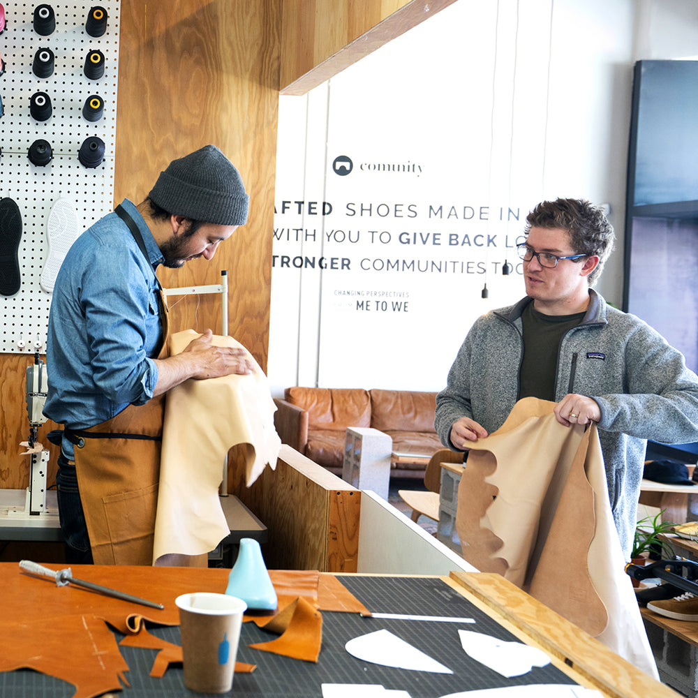 A photo of two men in a cobbler station surrounded by leather and shoe patterns who are working on making shoes