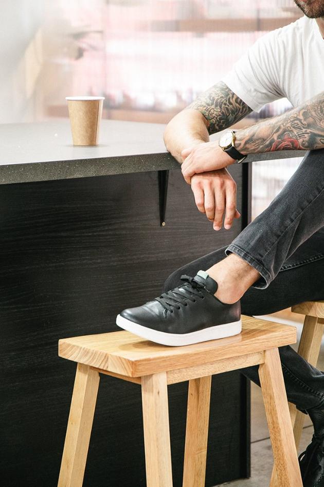 Man sitting on a stool with his foot propped up on another stool.  He is wearing a black leather sneaker with a white outsole