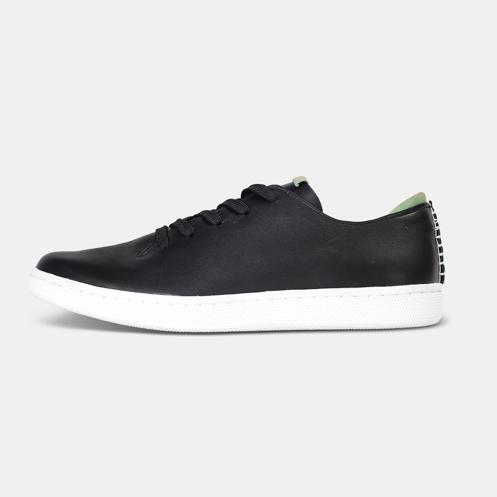 A profile shot of single black leather sneaker with a lace-up closure, featuring flat black shoelaces, a white rubber cupsole and a green accent on the heel.