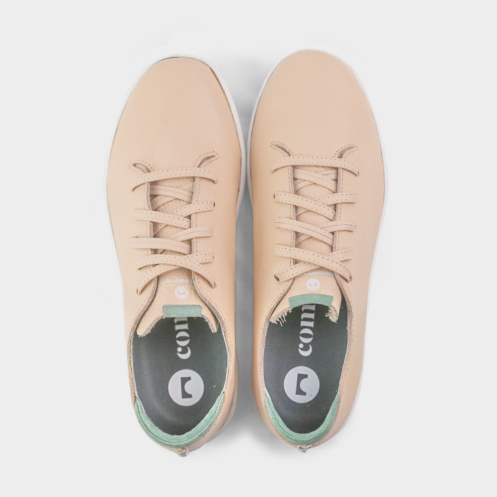 
                      
                        A top view shot of pair of nude beige leather sneakers with a lace-up closure featuring flat nude beige leather shoelaces, gold printing on the tongue and a dark grey sockliner with a circle logo and COMUNITY printed in white with a green heel accent.
                      
                    