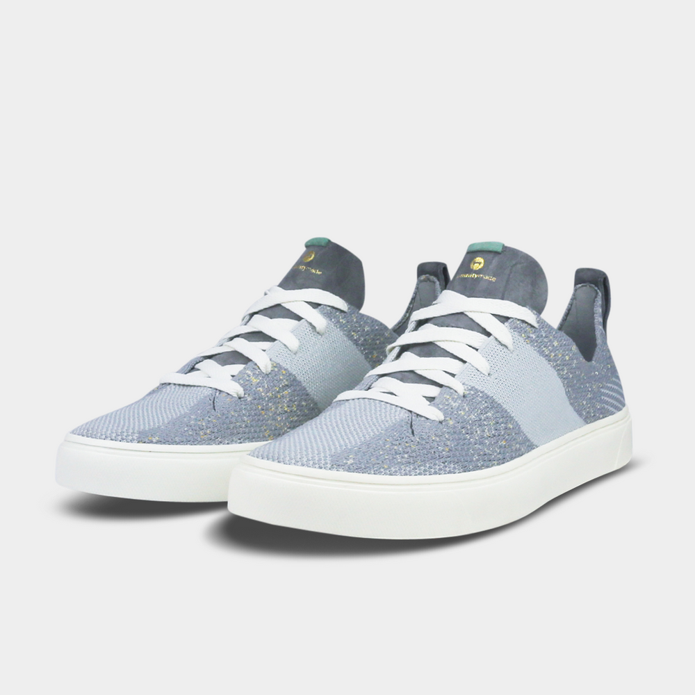 A pair of light gray knitted sneakers. The shoes have a thick, gray suede pull tab on the back with a gold logo and a small mint green detail at the base.