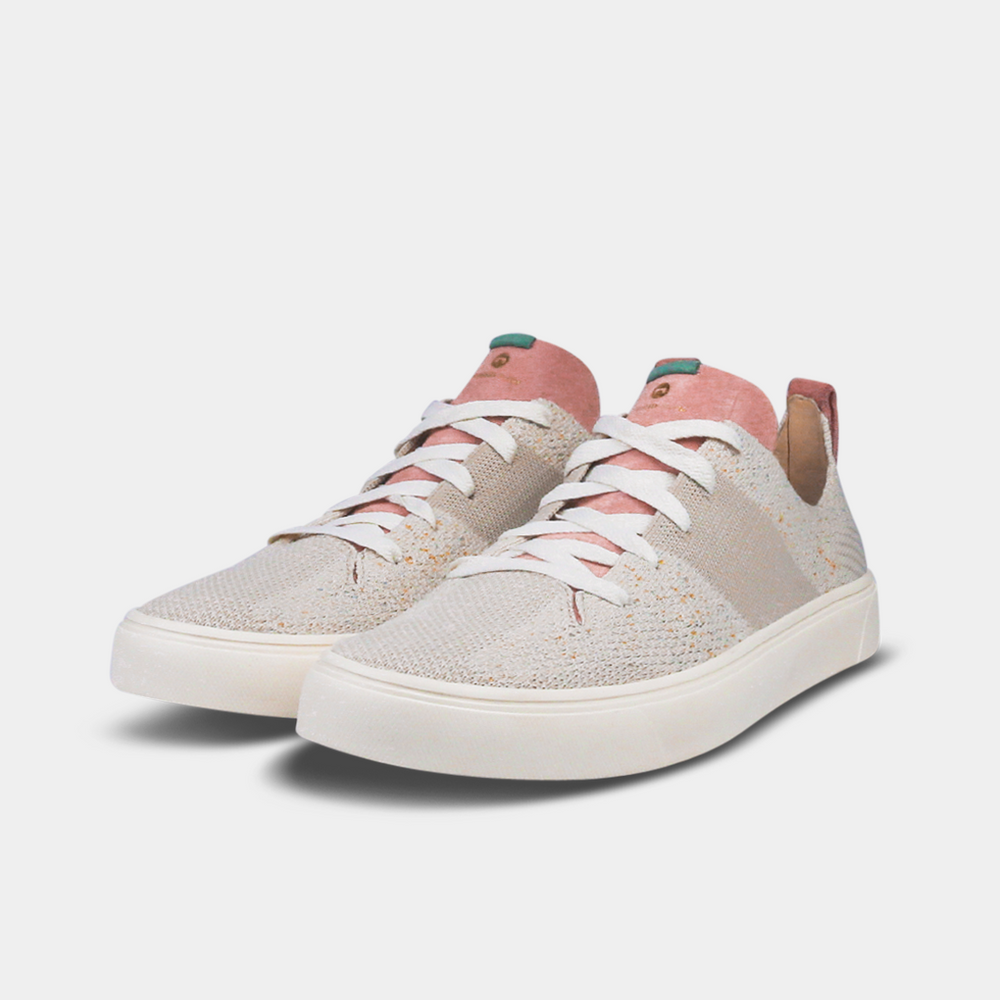 A pair of off-white knitted sneakers with light pink speckles. The shoes have white laces, a pale pink tongue, and a pink and white heel loop. The insoles are dark green with a white text logo and a circle.