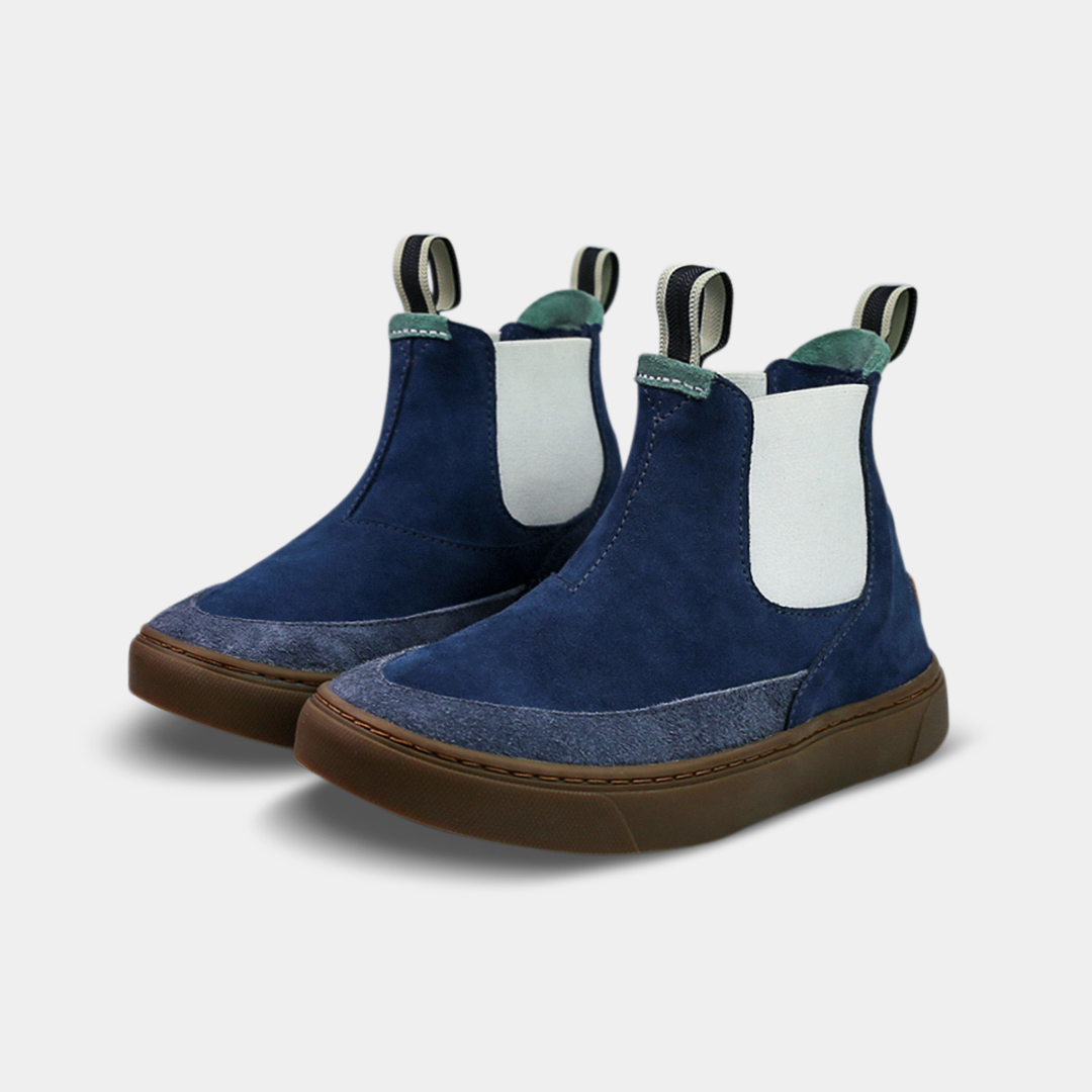 A pair of dark blue suede ankle boots with a brown rubber sole. The boots feature a light brown leather logo tag with "DEVIUM" written on the back of each heel, a mint green elasticated panel, and a black and white striped pull tab at the back of the ankle.