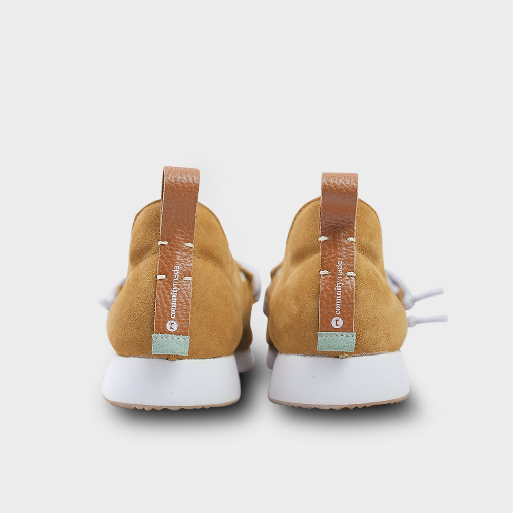 
                      
                        A back-angle, heel product shot of a pair of brown suede slip-on shoes. The shoes have thick, speckled black and white rope-like laces that are tied in a loose knot across the top of the foot. The insoles are dark gray with the word "comunity" and a circular logo printed on them in white. The shoes have a small brown leather loop on the heel. The background is a solid light gray.
                      
                    