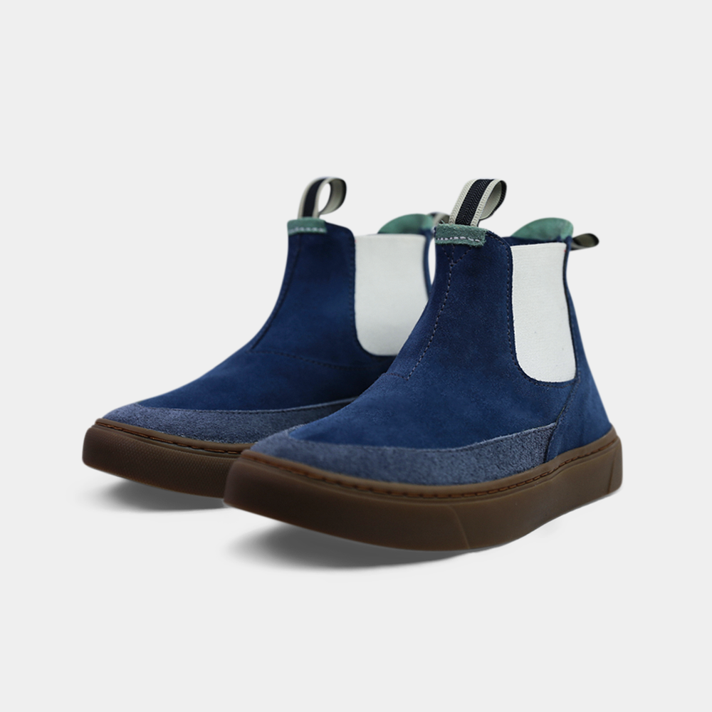 A pair of dark blue suede ankle boots with a brown rubber sole. The boots feature a light brown leather logo tag with "DEVIUM" written on the back of each heel, a mint green elasticated panel, and a black and white striped pull tab at the back of the ankle.