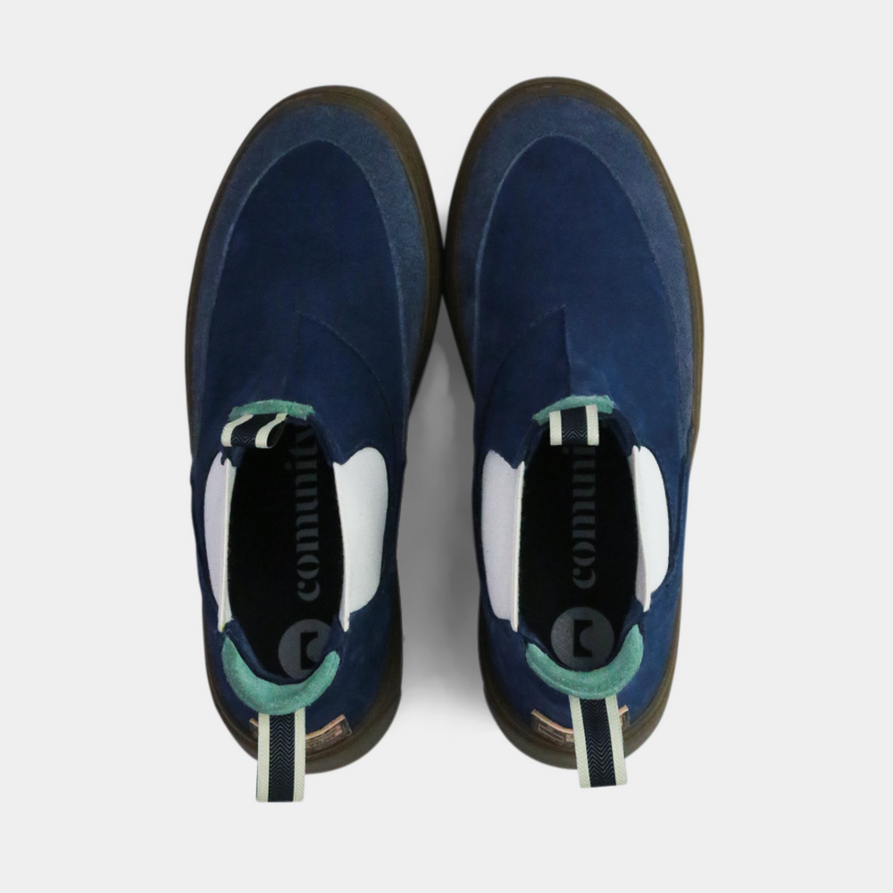 
                      
                        A pair of dark blue suede ankle boots with a brown rubber sole. The boots feature a light brown leather logo tag with "DEVIUM" written on the back of each heel, a mint green elasticated panel, and a black and white striped pull tab at the back of the ankle.
                      
                    