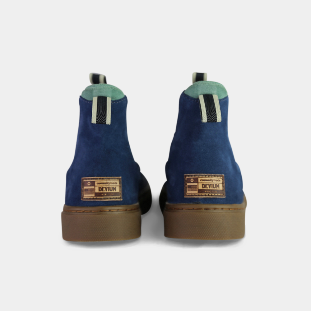 
                      
                        A pair of dark blue suede ankle boots with a brown rubber sole. The boots feature a light brown leather logo tag with "DEVIUM" written on the back of each heel, a mint green elasticated panel, and a black and white striped pull tab at the back of the ankle.
                      
                    
