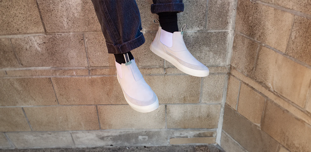 A pair of legs wearing black socks and dark, cuffed jeans is shown. A pair of off-white, high-top slip-on sneakers with white soles are on the feet. The Devium sneakers have a clean, minimalist design with a small green loop on the back of the ankle. The person is sitting on a ledge with their feet dangling in front of a brick wall.