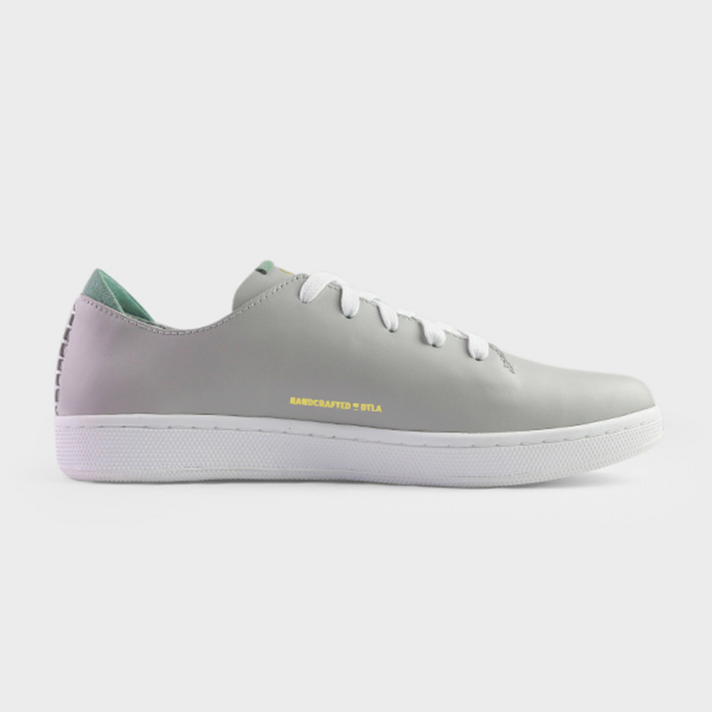 
                      
                        A medial profile shot of single light grey leather sneaker with a lace-up closure, featuring flat white shoelaces, white accent stitching on the heel, gold "Handcrafted in LA" printing on the instep and a white rubber cupsole.
                      
                    
