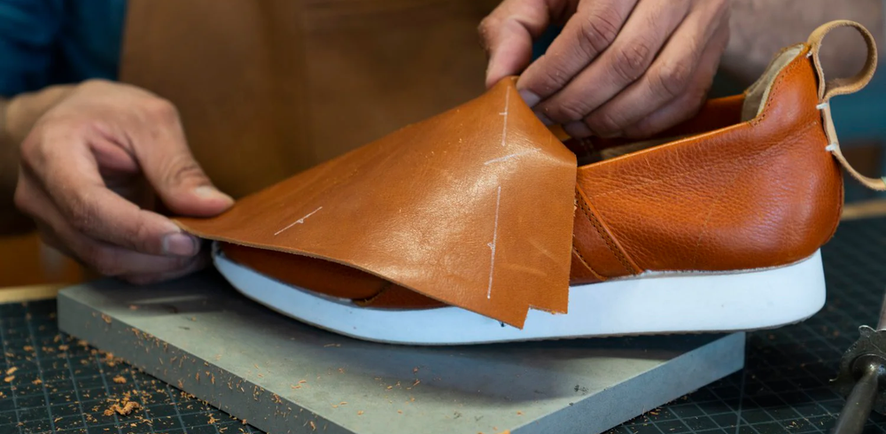 A close-up shot of a person's hands in a workshop, working on a shoe. The person is holding a light brown piece of leather, which has white marks on it, and is draping it over a partially completed orange-brown leather shoe. The shoe has a thick white sole, and the person's hands are carefully aligning the leather piece to the body of the shoe. The work is being done on a small gray mat.