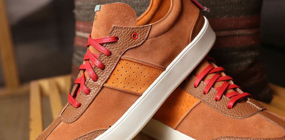 Close up shoe of a pair of brown suede shoes that have a lace up closure with red leather laces.  The bottom, of the shoe is a  white rubber cupsole