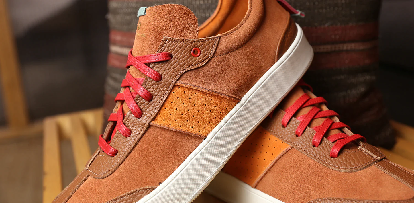 Close up shoe of a pair of brown suede shoes that have a lace up closure with red leather laces.  The bottom, of the shoe is a  white rubber cupsole